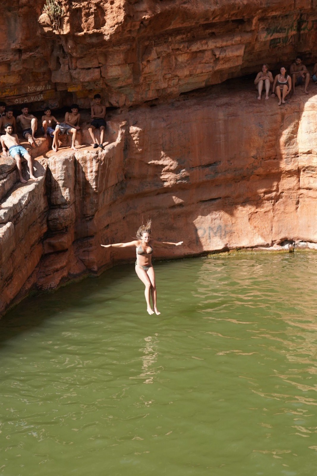 Mujer saltando desde las rocas a la piscina verde de Paradise Valley mientras otros miran