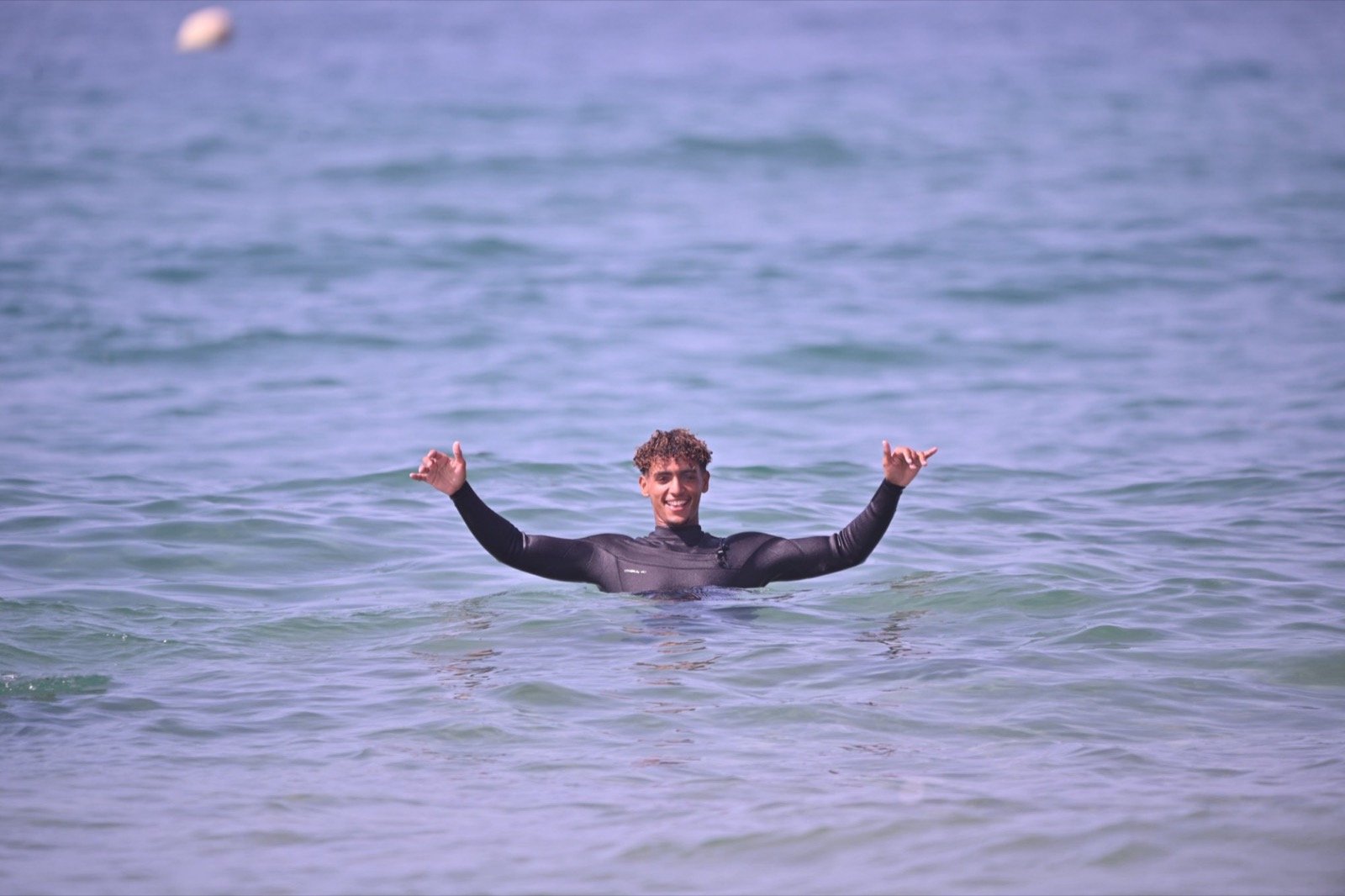 Ismail en el agua haciendo el signo shaka con las dos manos después de una clase de surf
