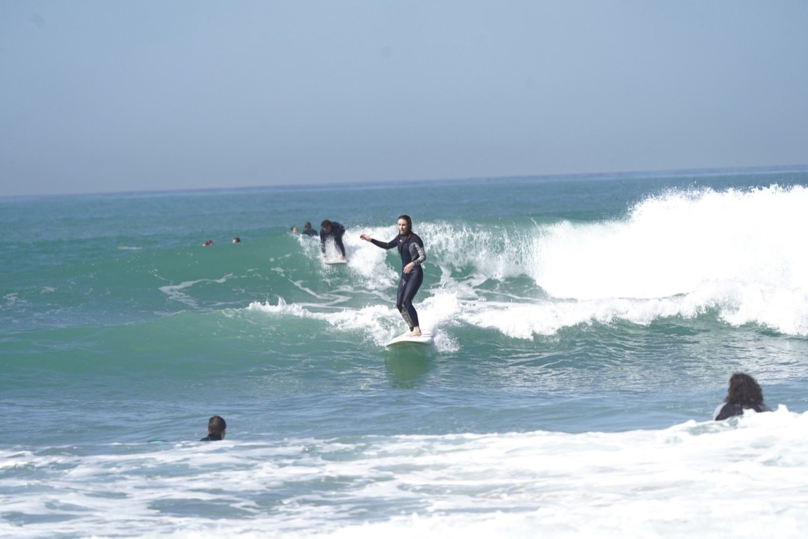 Alumno de surf surfeando una ola sobre una tabla blanca con otros surfistas alrededor en Taghazout