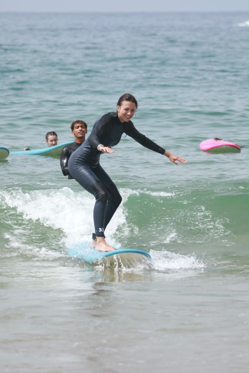 Surf student riding a wave in Taghazout with her coach behind her